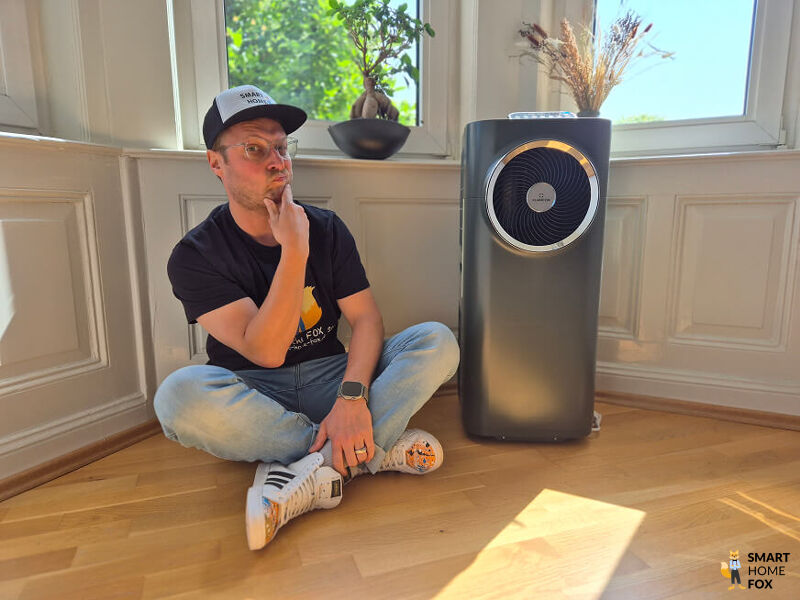 Smart-Home-Fox Experte sitzt im Test nachdenklich neben mobilem Klimagerät im Schlafzimmer